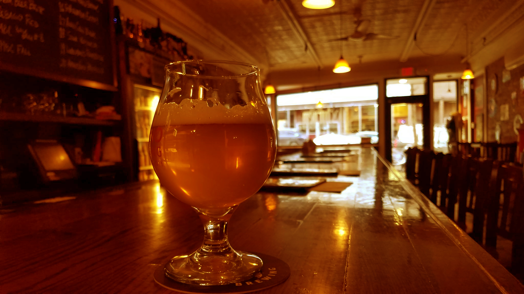 A golden beer in a snifter on the long wooden bar, front windows glowing at dusk.